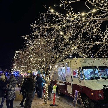 holiday lights wrapped around a tree with people buying food from a food truck in winter
